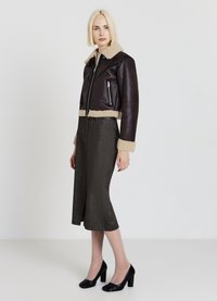 Giacca in pelle marrone, taglio corto, con colletto e polsini in shearling beige, tasca sul petto con zip, abbinata a una gonna midi a fantasia scura e tacchi neri.