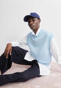 Lichtblauwe mouwloze trui over een wit overhemd met kraag, gecombineerd met marineblauwe broek met krijtstrepen. Model draagt een marineblauwe pet. Gladde texturen zichtbaar.