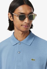 Világoskék poloing, texturált anyagból, három gombbal. Átlátszó napszemüveg sötét lencsékkel és Lacoste logóval a kereten.