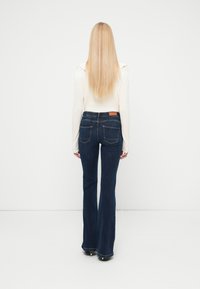 Γυναίκα με μακριά ξανθά μαλλιά που φοράει κρεμ πουλόβερ και σκούρα μπλε τζιν καμπάνα, στέκεται αντικρίζοντας έναν απλό λευκό τοίχο.