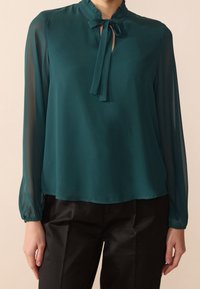 Blouse teal avec un col froncé haut et un détail de nœud, dotée de longues manches transparentes et d'une coupe ample, accompagnée de pantalons noirs.