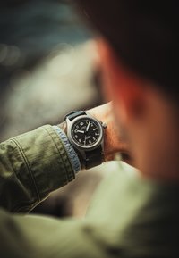Reloj analógico negro con caja redonda de acero inoxidable, grandes números blancos y correa de cuero negra. Lleva puesto en una muñeca con una camisa verde.