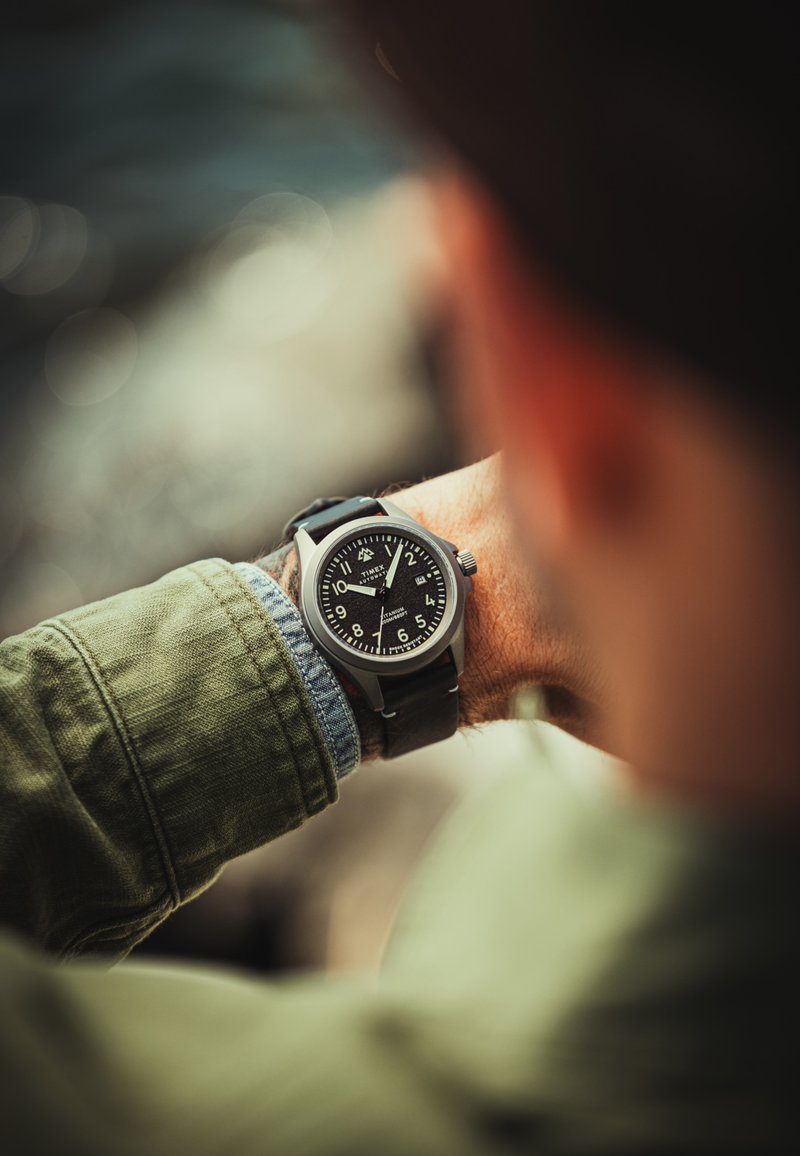 Reloj analógico negro con caja redonda de acero inoxidable, grandes números blancos y correa de cuero negra. Lleva puesto en una muñeca con una camisa verde.