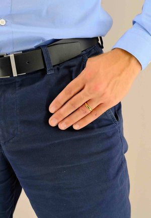 Fede nuziale in oro su una mano maschile che riposa su pantaloni blu navy, indossati con una camicia azzurra chiara e una cintura nera con fibbia argentata, mostrando texture lisce.