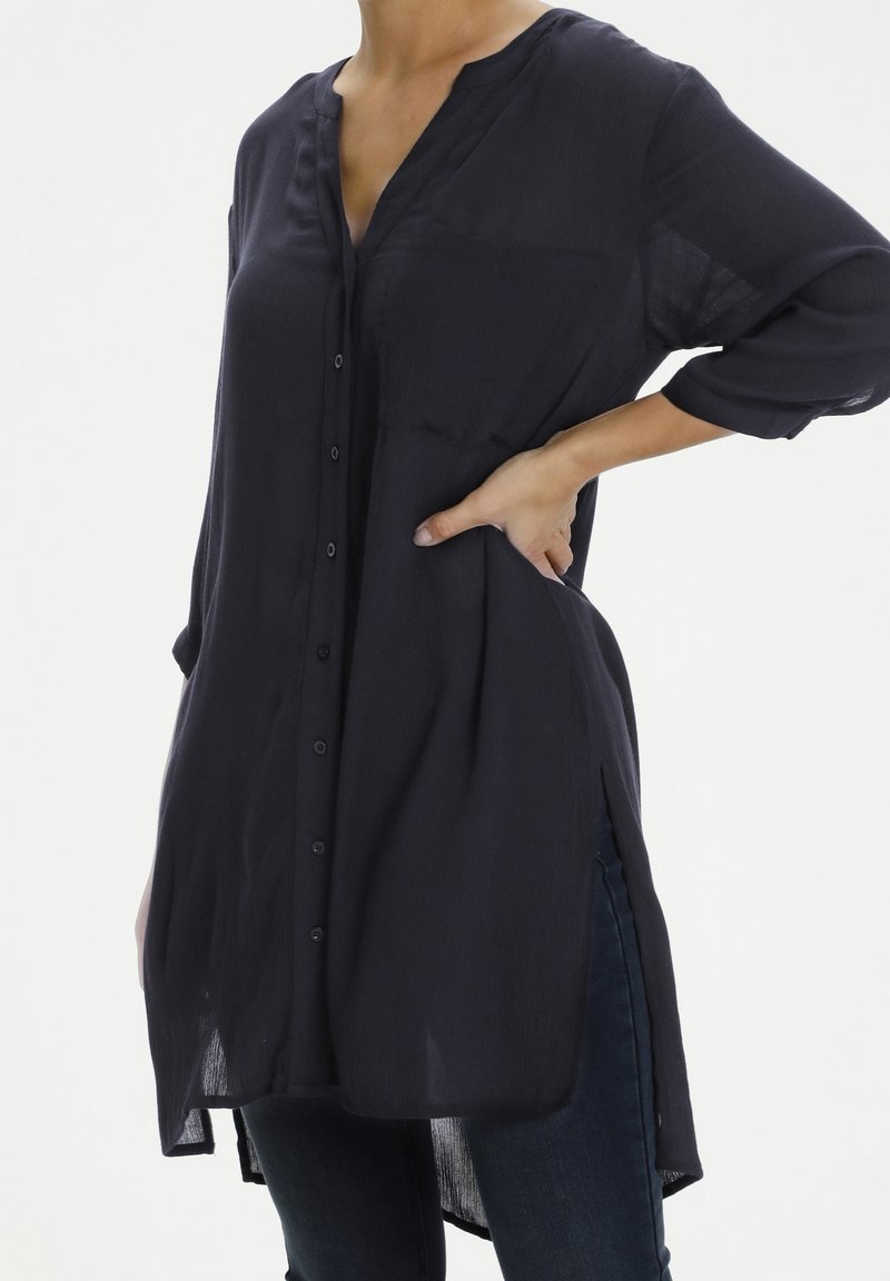 Chemise bleu marine à manches longues avec décolleté en V, devant boutonné et fentes sur les côtés. Confectionnée en tissu léger, avec deux poches poitrine.