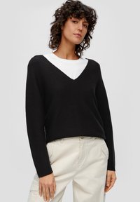 Schwarzer Strickpullover mit V-Ausschnitt, langen Ärmeln, gerippten Bündchen, über einem weißen Rundhals-Shirt getragen, kombiniert mit beigen Hosen.