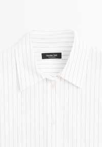 Bílá košile s tenkými námořnickými vertikálními pruhy, s rozloženým límečkem a přední légou. Etiketa zní "Massimo Dutti, vyrobeno v Maroku."