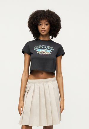 Νεαρή γυναίκα με σγουρά μαλλιά που φοράει μαύρο cropped μπλουζάκι Rip Curl και μπεζ πλισέ φούστα, στέκεται μπροστά σε απλό φόντο.