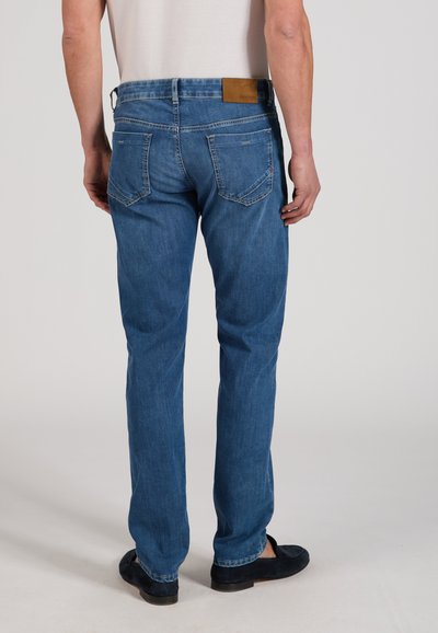 Man wearing blue denim jeans and black slip-on shoes, standing facing away against a plain light background.