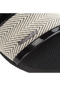 Musta-valkoinen Havaianas-liukusandaali, jossa on sahalaitakuosilla varustettu kangashihna ja teksturoitu kumipohja, jossa on sileä musta yksityiskohta.
