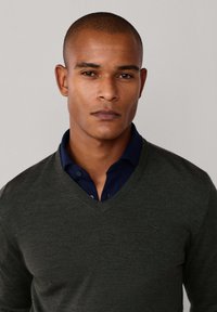 Donkergroen, lichtgewicht v-hals trui gelaagd over een marineblauw overhemd met kraag. Bevat een subtiel embleem op de linkerkant van de borst. Glad textuur.