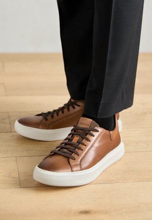 Bruine leren sneakers met een witte rubberen zool, voorzien van bruine veters en een logo-detail. Draagt met zwarte broek en een zwarte sok.