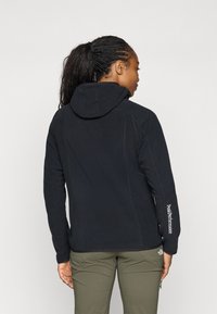 Zwarte fleecetrui met een capuchon, met gestikte details en een logo op de mouw. De persoon draagt licht olijfgroene broek.