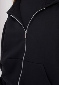 Acercamiento de una sudadera negra con cremallera y un bolsillo frontal, llevada sobre una camiseta negra.