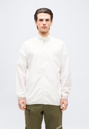 Junger Mann mit zurückgekämmtem Haar, trägt eine weiße Jacke mit Reißverschluss und olivgrüne Cargo-Hosen vor einfachem Hintergrund.