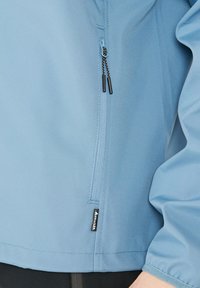 Ljusblå mjukskaljacka med en blixtlåsförsedd ficka, svarta dragkedjor och en märkesskylt vid fållen. Slät textur och skräddarsydd passform.