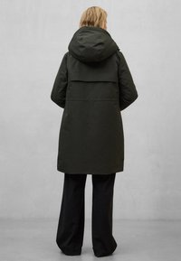 Tmavě zelená dlouhá bunda s kapucí, volného střihu a překládanou zadní klopou, doplněná černými širokými kalhotami v minimalistickém prostředí.