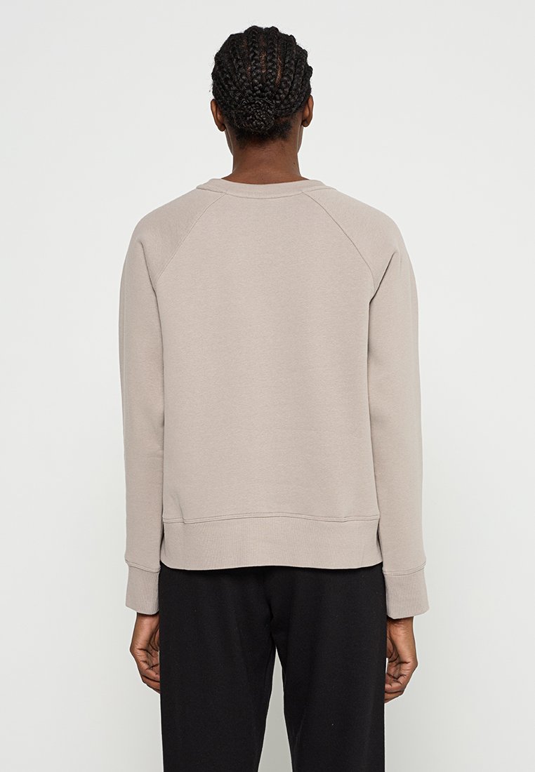 Osoba z warkoczami, ubrana w beżowy długi rękaw sweter i czarne spodnie, widziana od tyłu na jednolitym tle.