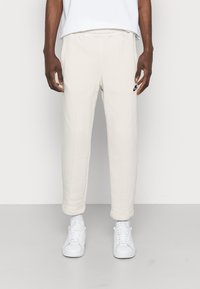 Beige svettbyxor med en avslappnad passform, med sidofickor och en liten svart logotyp på vänster sida, i kombination med vita sneakers.