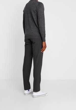 Person iført mørkegrå langærmet sweater, mørkegrå bukser og hvide højtop-sneakers, stående med ryggen mod en ensfarvet hvid baggrund.
