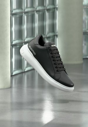 Μαύρο αθλητικό παπούτσι Valentino με λευκή σόλα που αιωρείται πάνω από ένα γυαλισμένο σκυρόδεμα σε μοντέρνο εσωτερικό χώρο με τοίχους από γυάλινους κύβους.