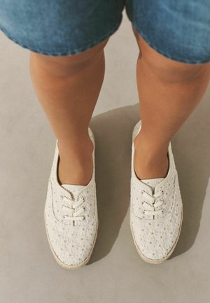 Osoba nosící bílé květinové šněrovací boty a modré džínové šortky, stojící na světle šedé podlaze.