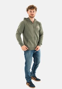 Sweat à capuche zippé vert avec poches avant, associé à un jean bleu et des sneakers noires avec des accents marron. Design décontracté et coupe relax.