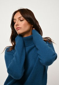 Tyrkis turtlenecktröja med en mjuk textur, avslappnad passform och ribbade manschetter, bärs med händerna som kupa halsringningen.