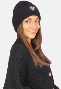 Bonnet en tricot noir avec texture en côtes et patch en forme de cœur en imprimé léopard. Modèle portant un cardigan en tricot noir assorti.