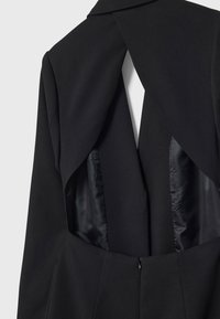 Vêtement noir à manches longues avec un dos ouvert dévoilant une doublure en satin noir à l'intérieur de la découpe.