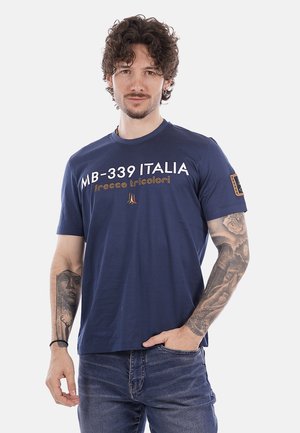 Hombre con pelo rizado y tatuajes que lleva una camiseta azul marino "MB-339 ITALIA frecce tricolori" y vaqueros azules, de pie con una mano en el bolsillo.