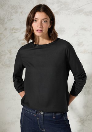 Zwarte lange mouw top met een ronde halslijn en gestippelde afwerking. Soepele stof en een relaxte pasvorm. Gecombineerd met donkerblauwe jeans.