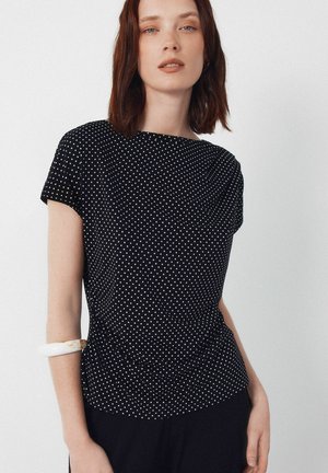 Femme aux cheveux bruns mi-longs portant un haut noir à manches courtes à pois blancs et un bracelet blanc sur le bras gauche.