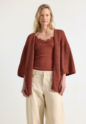 Donna bionda che indossa una camisole color ruggine con bordatura in pizzo e un cardigan lavorato a maglia abbinato con pantaloni crema a gamba larga, in piedi su sfondo bianco.
