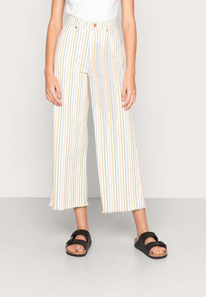 Personne portant un pantalon blanc à rayures multicolores verticales et des sandales noires à deux brides, debout devant un fond clair uni.