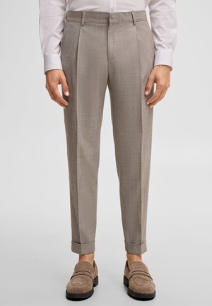 Mężczyzna w beżowych spodniach szytych na miarę z przednimi plisami, podwiniętymi nogawkami, w połączeniu z mokasynami ze skóry zamszowej i białą koszulą wizytową.