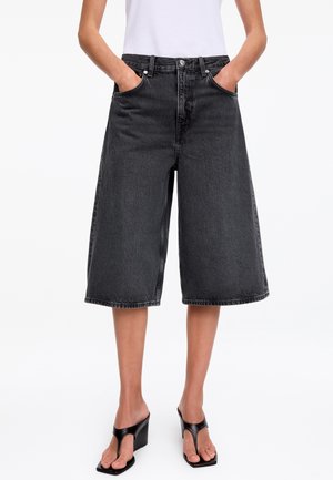 Personne portant un culotte en denim noir taille haute avec poches, associée à des sandales à talons noires et un haut blanc rentré.