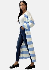 Lange, offene Strickjacke aus weichem Material mit blauen und weißen Streifen. Verfügt über ein geripptes Muster und weite Ärmel. Kombiniert mit eng anliegenden Jeans.