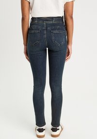 Helle Jeans aus dunkelblauem Denim mit einer hohen Taille, Gesäßtaschen und Stitching-Details. Kombiniert mit weißen Sneakers mit schwarzen Akzenten.