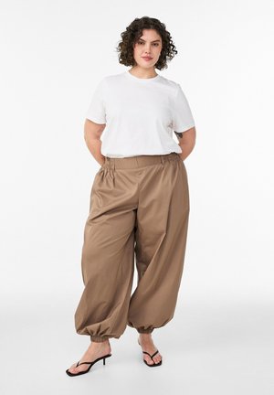 Žena kovrčave kose u bijeloj majici s kratkim rukavima, širokim smeđim hlačama s elastičnim manšetama i crnim sandalama na petu, stoji s rukama iza leđa.