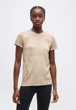 Nuori henkilö, jolla on punotut hiukset, beige lyhythihainen urheilupaita ja mustat leggingsit, seisoo yksivärisen taustan edessä.