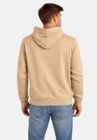 Uomo con felpa beige con cappuccio e jeans blu, in piedi di spalle contro uno sfondo chiaro e uniforme.