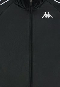 Crna zip-up jakna od glatkog materijala, s visokim okovratnikom i bijelim logotipom. Vertikalni zatvarač se proteže niz središnji dio. Bijeli akcenti na ramenima.