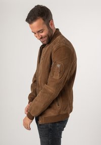 Giubbotto bomber in suede marrone con zip frontale, polsini a coste e tasca a toppa sul braccio sinistro. Caratterizzato da una texture liscia e un design aderente.