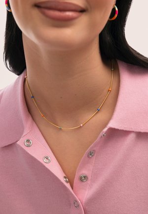Primer plano de una persona que lleva un delicado collar de oro con pequeñas cuentas multicolores y una camisa rosa con cuello y botones a presión plateados.