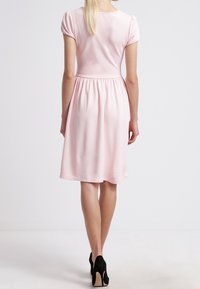 Femme aux longs cheveux blonds portant une robe rose clair à manches courtes arrivant au genou et des chaussures à talons hauts noires, se tenant de dos.