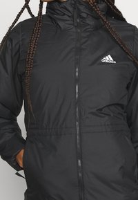 Giacca imbottita nera con alto colletto, zip frontale e polsini elasticizzati. Presenta un logo Adidas bianco sul petto. Tessuto liscio e leggero.