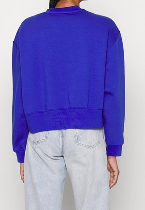 Personne vue de dos portant un sweat-shirt manches longues bleu roi et un jean taille haute bleu clair avec poches arrière.