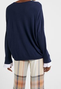 Granatowy, luźny sweter z szerokimi rękawami, z białymi plisowanymi akcentami przy mankietach, w zestawieniu z beżowymi spodniami w kratę.