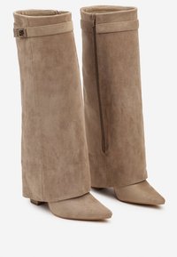 Bottes de cheville en suède beige au design décontracté, à bout pointu et à petit talon bloc. Présentent une fermeture éclair sur le côté et un accent de sangle au niveau du bracelet.
